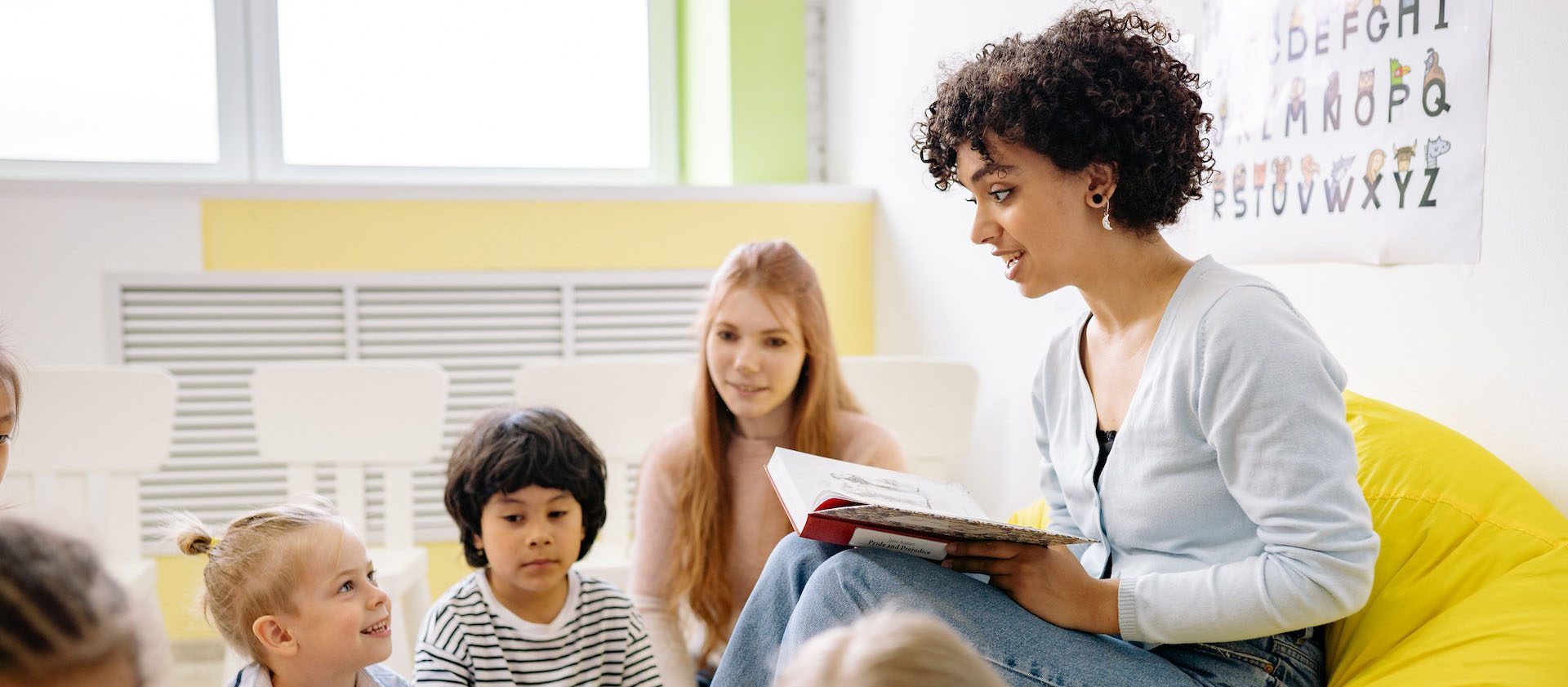 Teacher with curly hair reaching a book to students while sitting in a yellow bean bag chair.